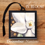 Framed artwork of white flowers with a black ribbon on a wooden surface, featuring '1 Year in Bloom' and 'December Narcissus'.
