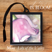 Framed artwork of a pink flower with 'May Lily of the Valley' text on a wooden background.