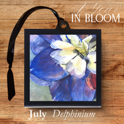 Framed artwork of a delphinium flower with a black ribbon on a wooden surface, titled '1 Year in Bloom'.
