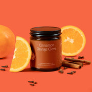 Candle jar labeled 'Cinnamon Orange Clove' surrounded by oranges and cinnamon sticks on an orange background