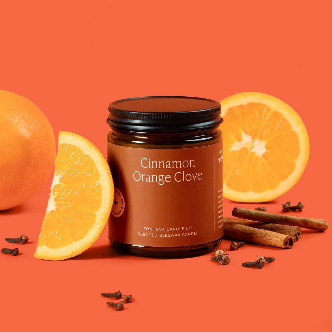 Candle jar labeled 'Cinnamon Orange Clove' surrounded by oranges and cinnamon sticks on an orange background