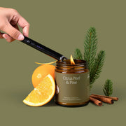 Hand lighting a candle labeled 'Citrus Peel & Pine' with oranges, cinnamon sticks, and pine branches on a green background.
