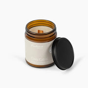 An amber glass jar candle with a cream label reading 'Fontana Candle Co.' and 'Cozy Cabin,' shown with its black lid resting beside it on a clean white background.