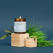 Candle labeled 'Cypress Sage & Patchouli' on a wooden block with green leaves against a blue background