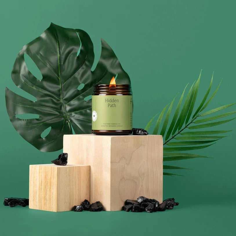 A lit candle in a dark amber glass jar with a light green label reading 'Hidden Path' sits on stacked light wood blocks against a teal-green background, styled with large green tropical leaves and scattered dark dried botanical pieces.