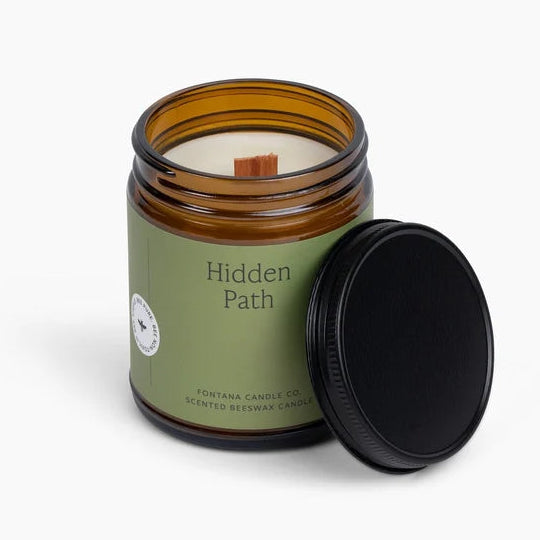 An amber glass jar candle with a sage green label reading 'Hidden Path' and a black lid resting beside it on a clean white background.