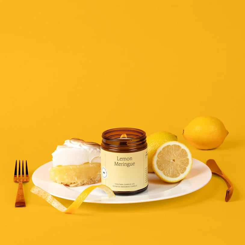 A candle in an amber glass jar labeled 'Lemon Meringue' sits on a white plate with a slice of lemon pie, a lemon half, whole lemons, a gold fork, and a gold spoon against a solid yellow background.