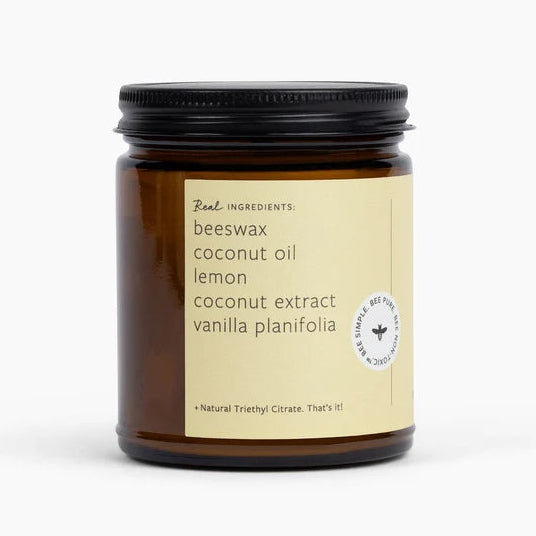 An amber glass jar candle with a black metal lid and a beige label. The label reads 'Real Ingredients beeswax coconut oil lemon coconut extract vanilla planifolia' on a clean white background.