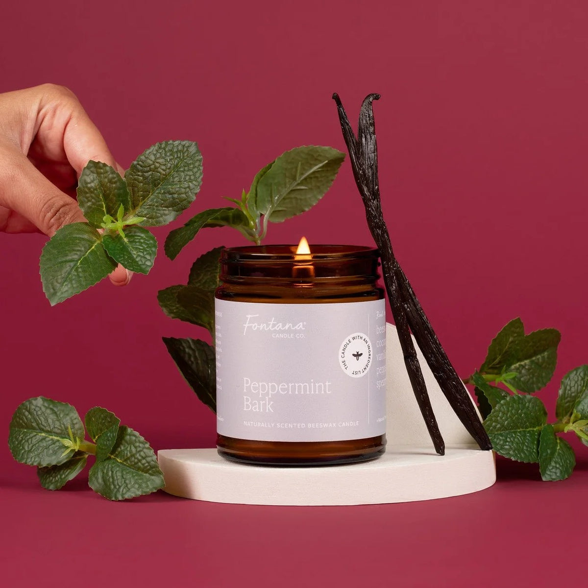 An amber glass jar candle labeled Fontana Scents Peppermint Bark, lit and styled with mint leaves and vanilla beans on a white pedestal against a dusty rose background.