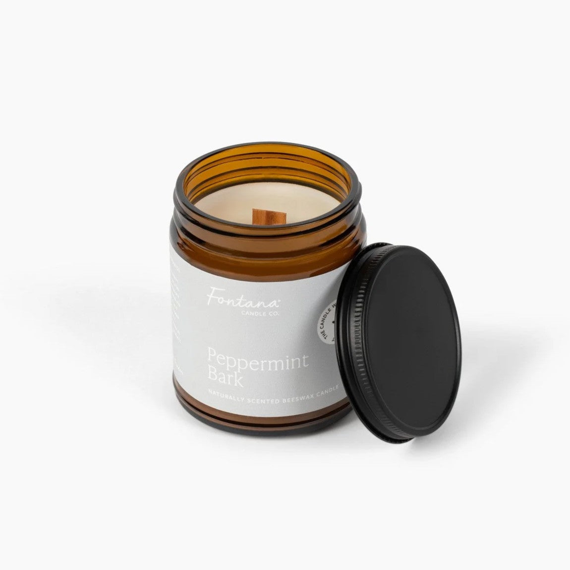 An amber glass jar candle with a white label reading 'Fontana Candle Co' and 'Peppermint Bark', shown with its black lid leaning against the jar on a clean white background.