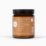 An amber glass jar candle with a black lid and a warm brown label, featuring text for 'Beeswax Coconut Oil Vanilla Planifolia Cinnamon Nutmeg Clove' on a clean white background.