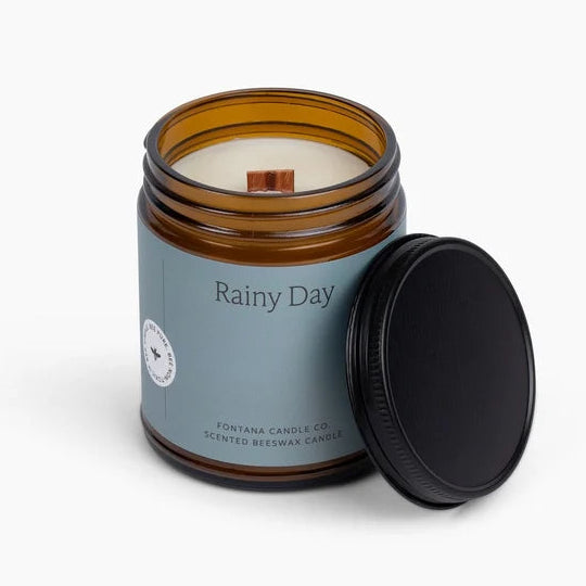 An amber glass jar candle with a light blue label reading 'Rainy Day' and a black screw-on lid leaning against the jar, photographed on a clean white background.