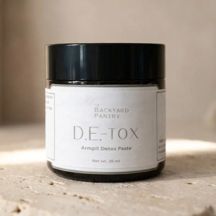 A jar of D.E-Tox Armpit Detox Paste by Backyard Pantry, featuring a black lid and a white label, set on a textured stone surface with a soft-focus beige background.