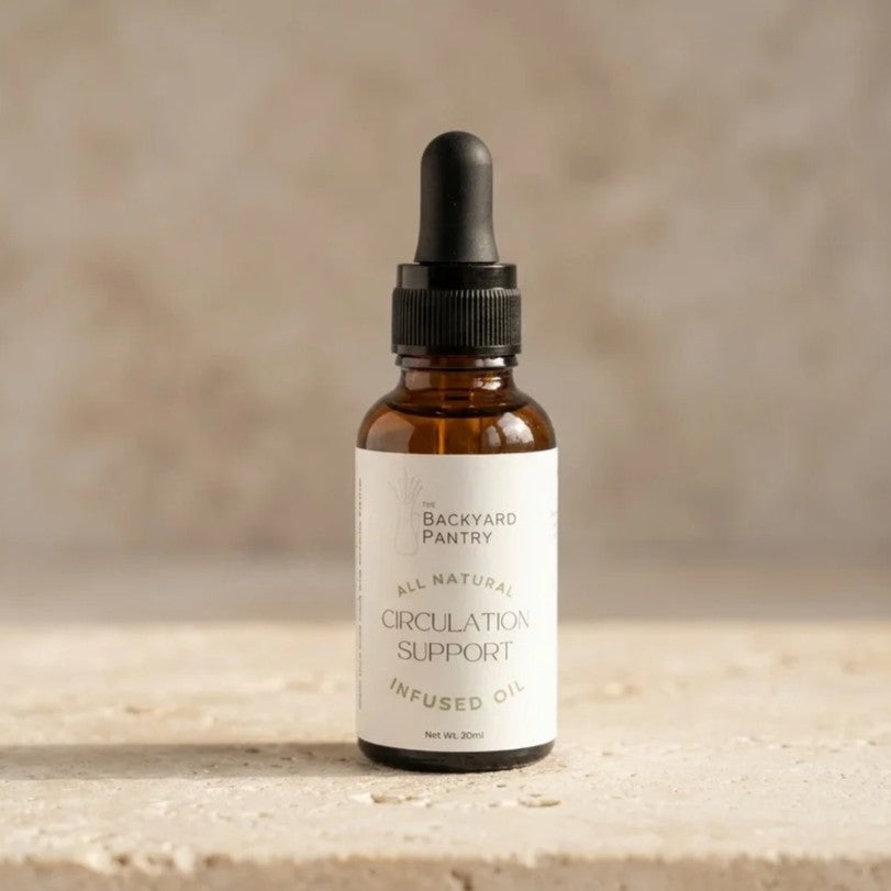 An amber glass dropper bottle of backyard pantry circulation support infused oil, standing on a textured stone surface against a neutral beige background.