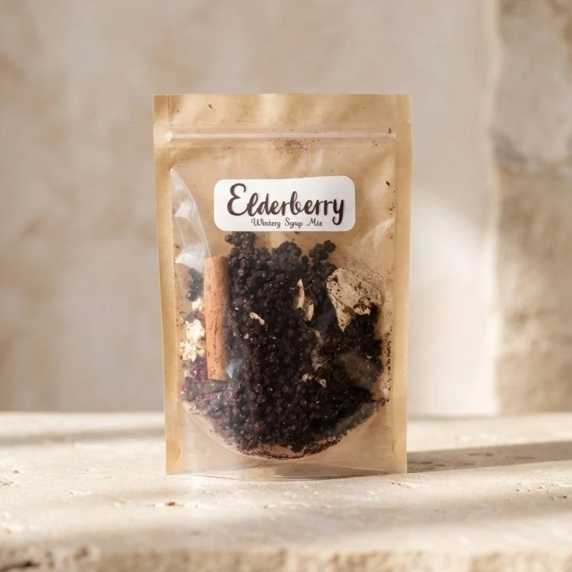 A stand-up pouch of Elderberry Wintery Syrup Mix containing dark elderberries, a cinnamon stick, and other spices, set against a rustic stone background.