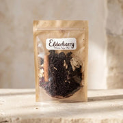 A stand-up pouch of Elderberry Wintery Syrup Mix containing dark elderberries, a cinnamon stick, and other spices, set against a rustic stone background.
