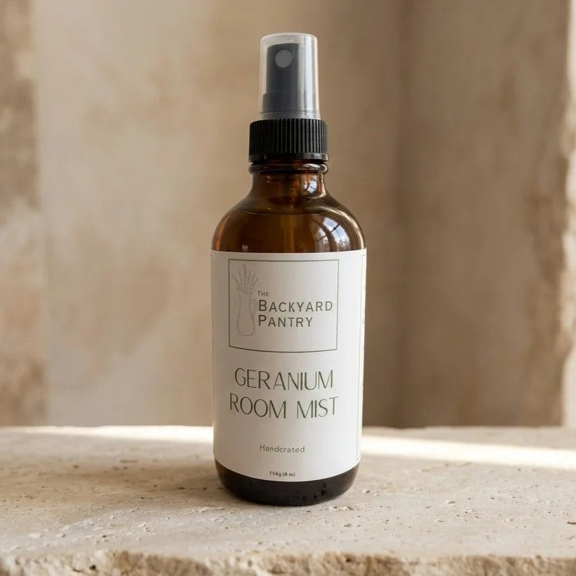 An amber glass room spray bottle with a white label reading 'The Backyard Pantry Geranium Room Mist' and a black spray top, set on a stone surface against a warm beige background.