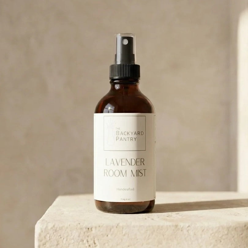 An amber glass room spray bottle with a black spray top and a cream label reading 'The Backyard Pantry Lavender Room Mist', set on a stone pedestal against a textured beige wall.