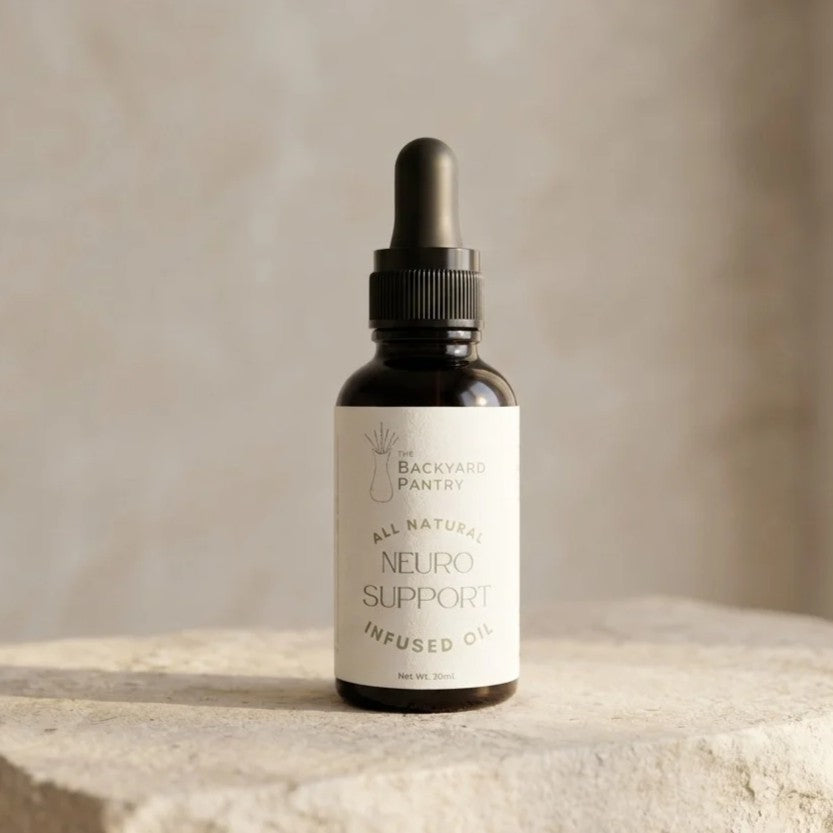 A small amber glass dropper bottle with a black cap and a cream label reading 'The Backyard Pantry' and 'All Natural Neuro Support Infused Oil,' set on a stone surface against a soft beige background.