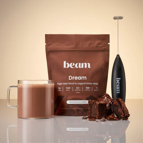 beam Dream chocolate product with a cup of hot chocolate, brownies, and an electric frother on a reflective surface.