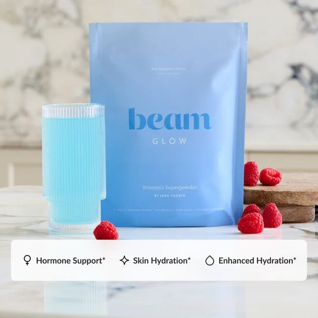 A light blue stand-up pouch labeled 'beam GLOW' stands on a marble surface beside a matching light blue drink in a ribbed glass, with raspberries placed around it. A white banner at the bottom lists benefits: Hormone Support, Skin Hydration, and Enhanced Hydration.