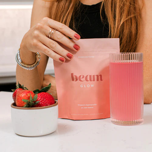 Person holding a 'beam Glow' package with a glass of pink drink and strawberries on a table.