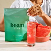 Person holding a green 'beam' package with a glass of watermelon juice and watermelon slices on a wooden table.