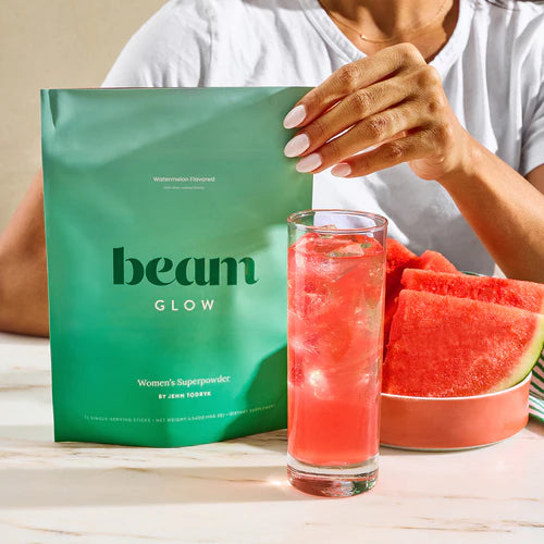 Person holding a green 'beam' package with a glass of watermelon juice and watermelon slices on a wooden table.