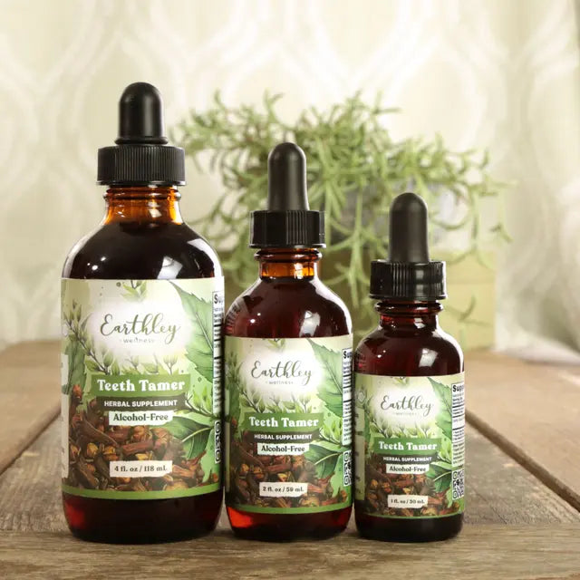 Three bottles of Earthley Herbals Teeth Tamer on a wooden surface with a blurred green plant background.